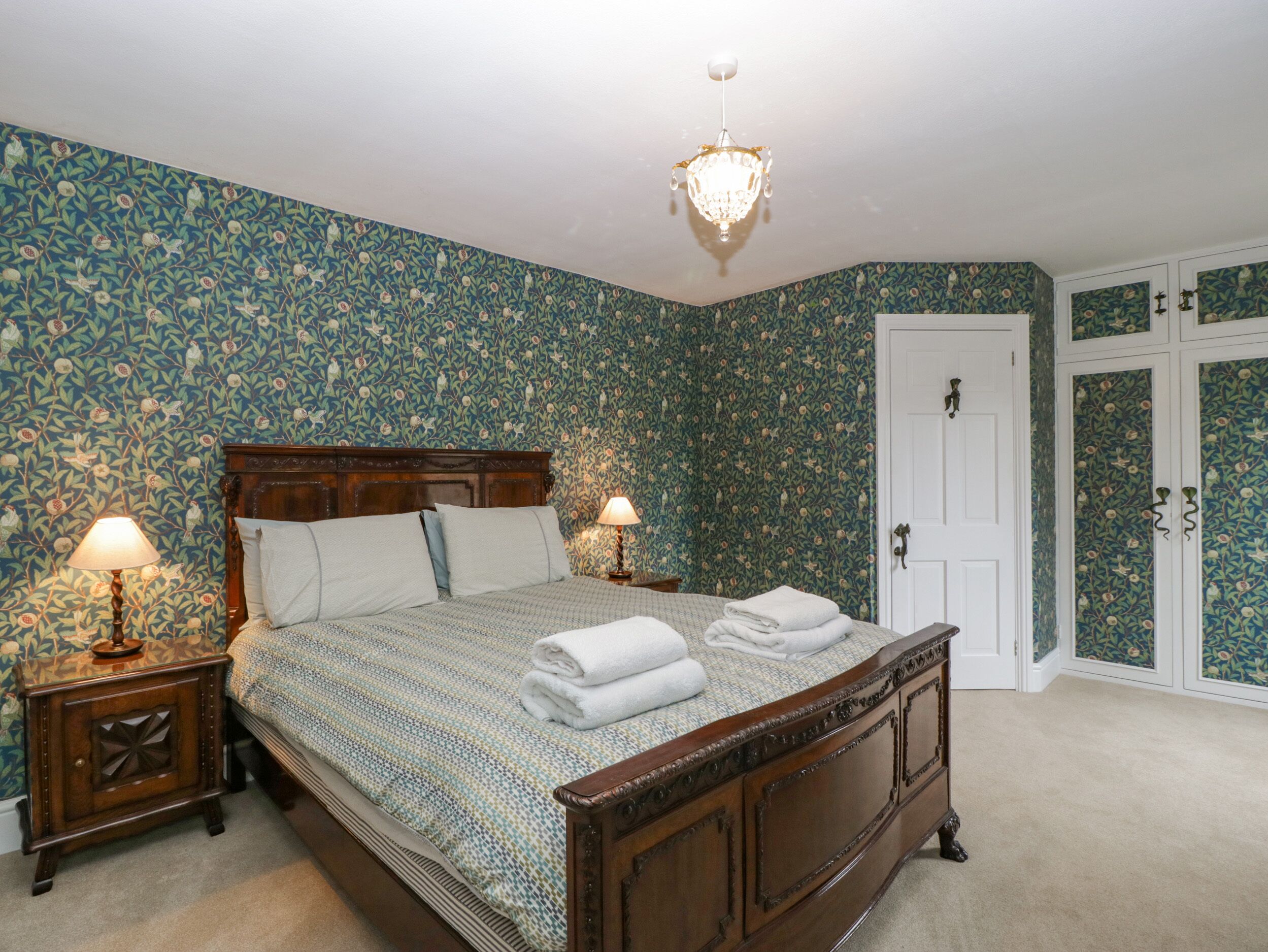 Un dormitorio con papel tapiz floral, una cama de madera con dosel, dos mesitas de noche con lámparas y un armario con estampado floral.