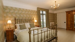 Un dormitorio tradicional con cama de dos plazas, dos lámparas de noche, papel tapiz floral, un candelabro y vistas a través de una ventana con cortinas.