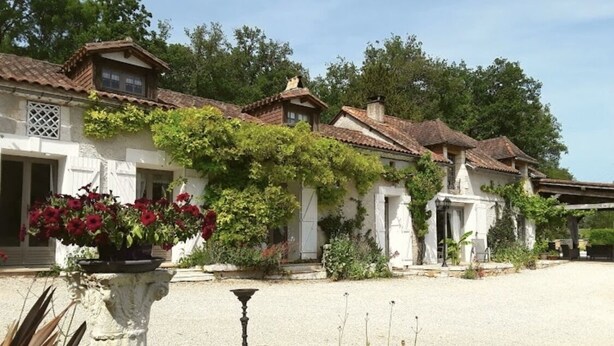 Une rangée de bâtiments traditionnels aux murs blancs et aux toits de tuiles, entourés de verdure et bordés d’une allée gravillonnée.