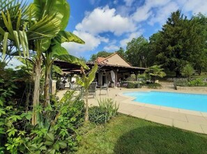 A pool area with a covered patio, lush greenery, and a house with a tiled roof.