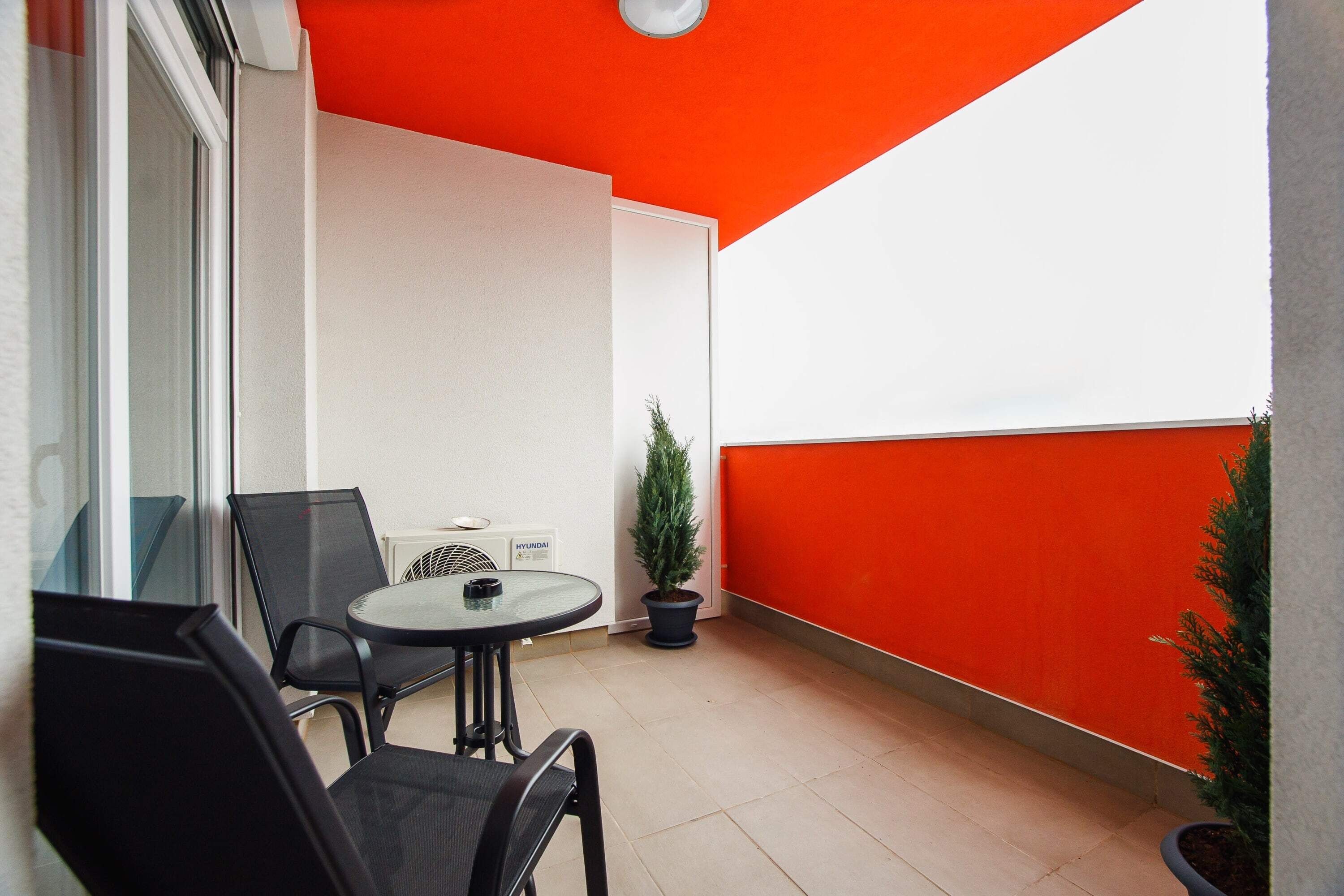 Un balcon avec une table ronde et deux chaises, un pot de fleurs et un mur rouge.