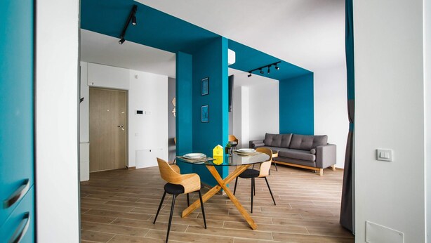 A modern living space with a dining area featuring a glass table and wooden chairs, a gray sofa, and a teal accent wall. - Azur Paradise near City Center (Oradea)