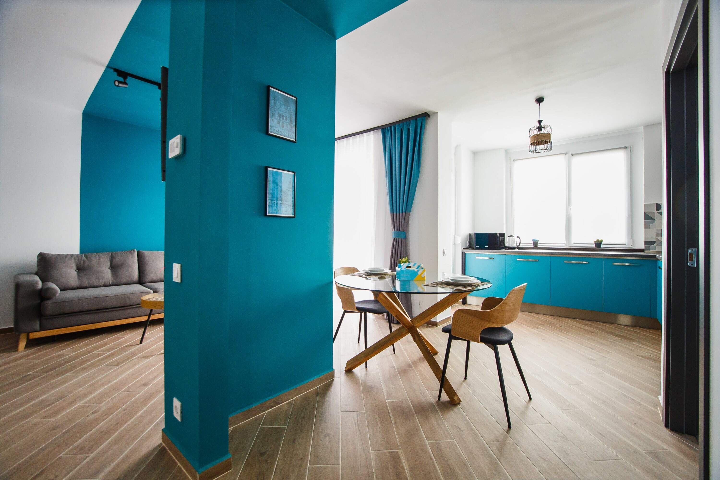 A modern living room with a sofa, a dining table with chairs, and a kitchen area with blue cabinets and a window.