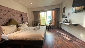 A hotel room with a large bed, a desk with a chair, a window with curtains, and a wall-mounted shelf. - The Saga Hotel (New Delhi)