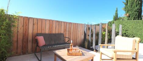 An outdoor patio with a wooden table, two chairs, a bench, and a bottle of wine.