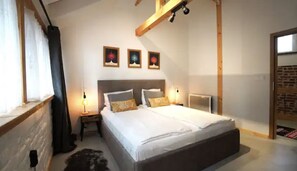 Un dormitorio con una cama grande, dos mesitas de noche y un techo inclinado con vigas a la vista. Hay tres cuadros enmarcados en la pared sobre la cama.
