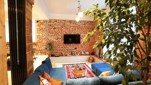 Una sala de estar con pared de ladrillo, un televisor de pantalla plana sobre un sofá y una planta grande en una esquina.