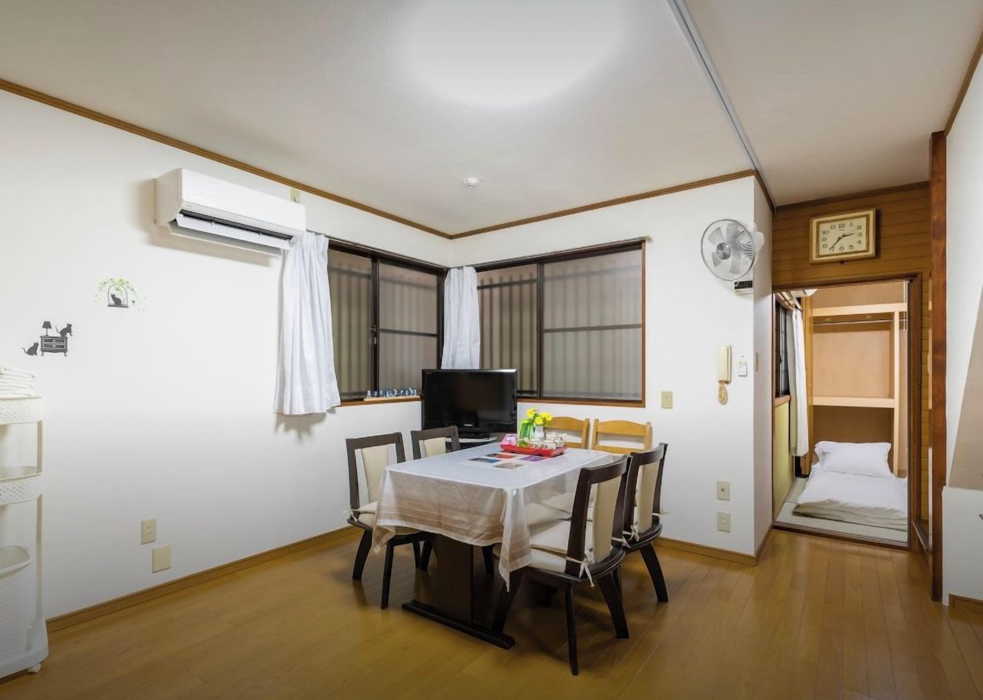 A room with a dining table, chairs, a TV, an air conditioner, and a bed.