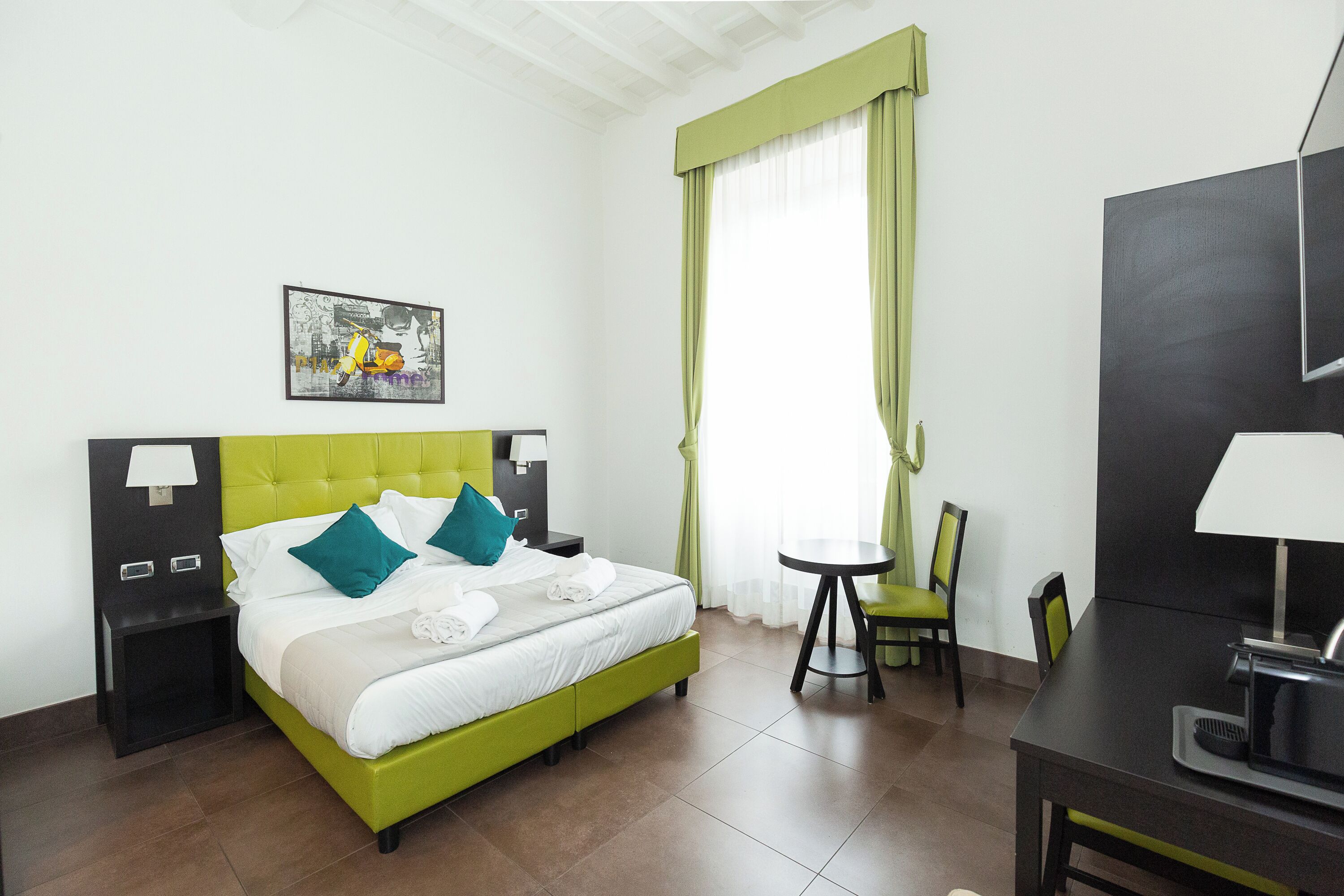 Habitación de hotel con cama verde, ropa de cama blanca, una mesita, una silla, un escritorio con lámpara y un cuadro en la pared.