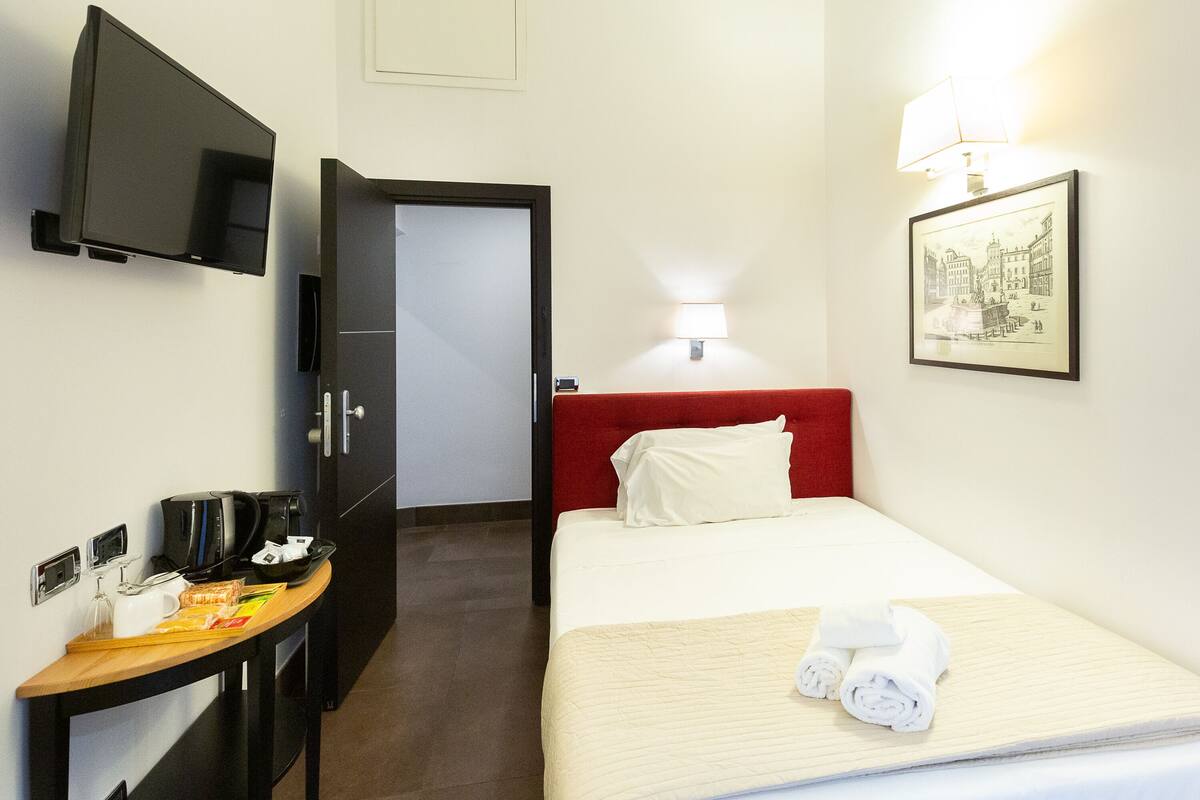 A hotel room with a bed, a desk with a coffee maker, a TV, and a framed picture on the wall.