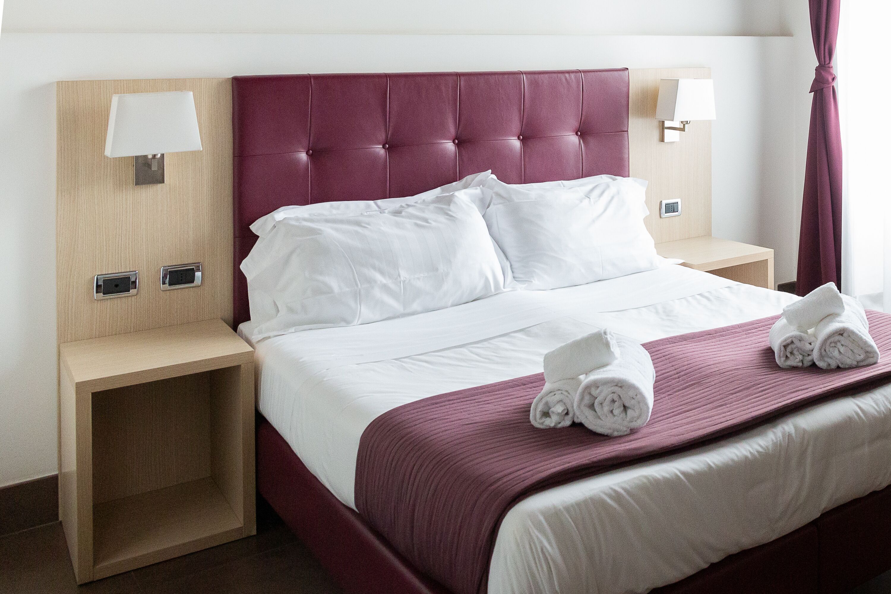 A neatly made bed with white linens and a burgundy throw blanket, two bedside tables with lamps, and a modern headboard.