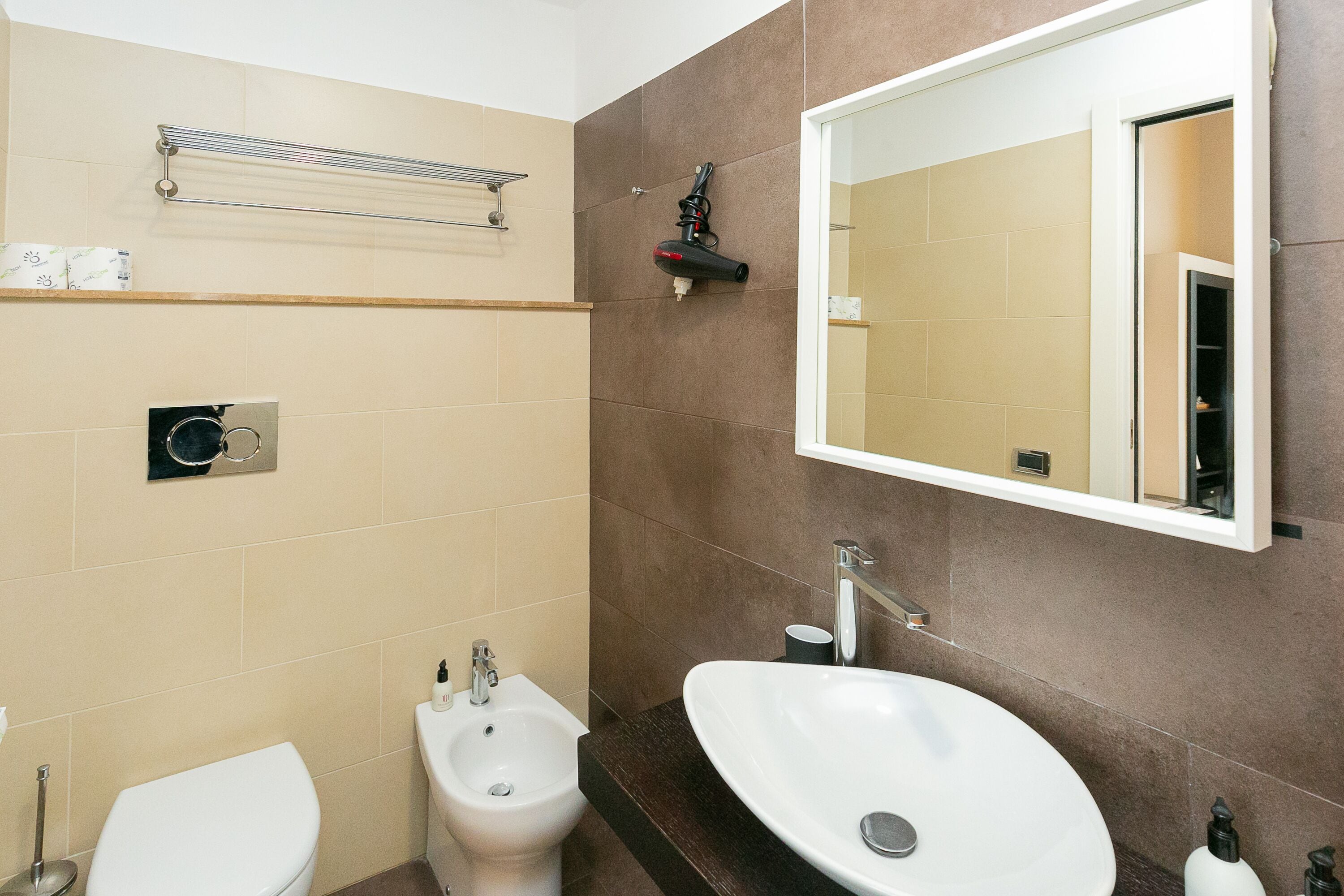 A bathroom with a toilet, bidet, sink, mirror, towel rack, and a shelf with toiletries.