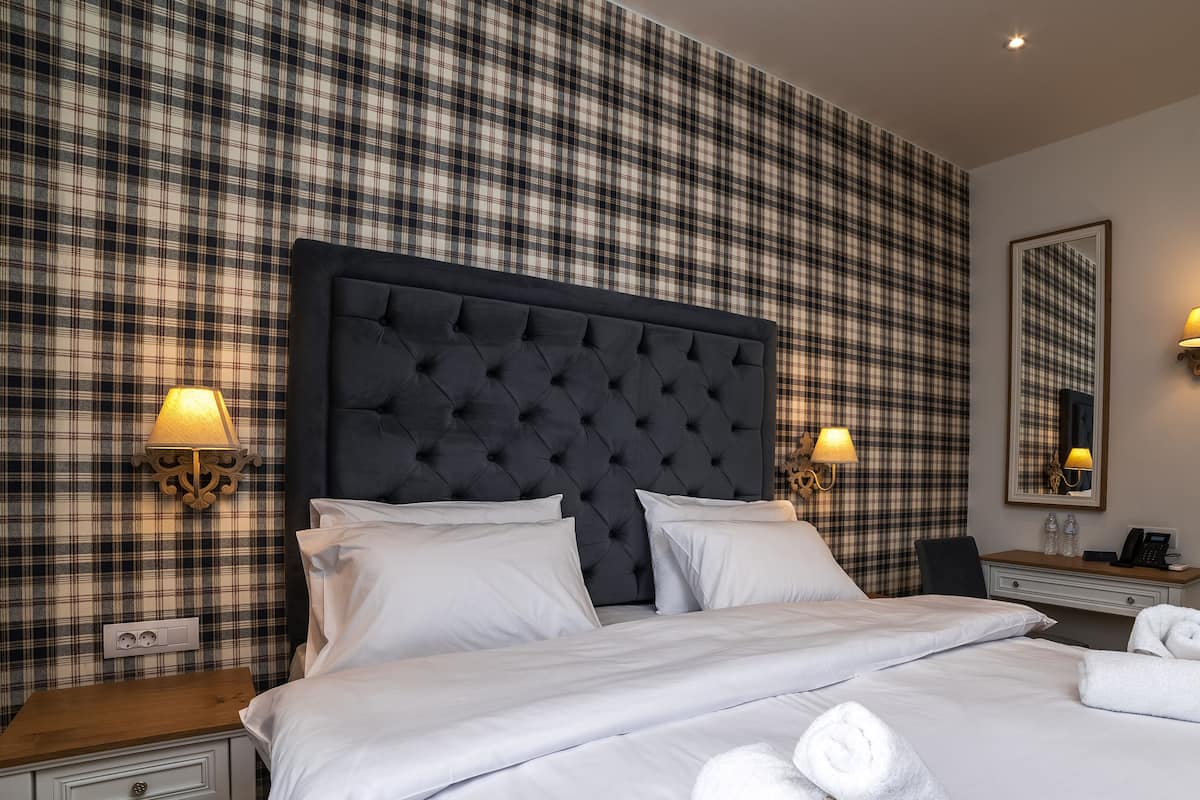 A hotel room with a plaid wallpaper, a tufted headboard, two bedside lamps, a bedside table with a phone and water bottles, and neatly folded towels on the bed.