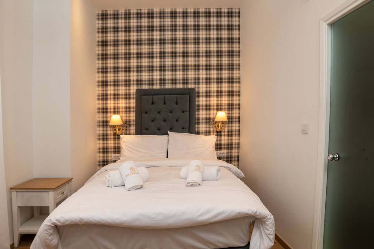 A neatly made bed with white linens and two pillows, a plaid-patterned headboard, and two wall-mounted lamps.
