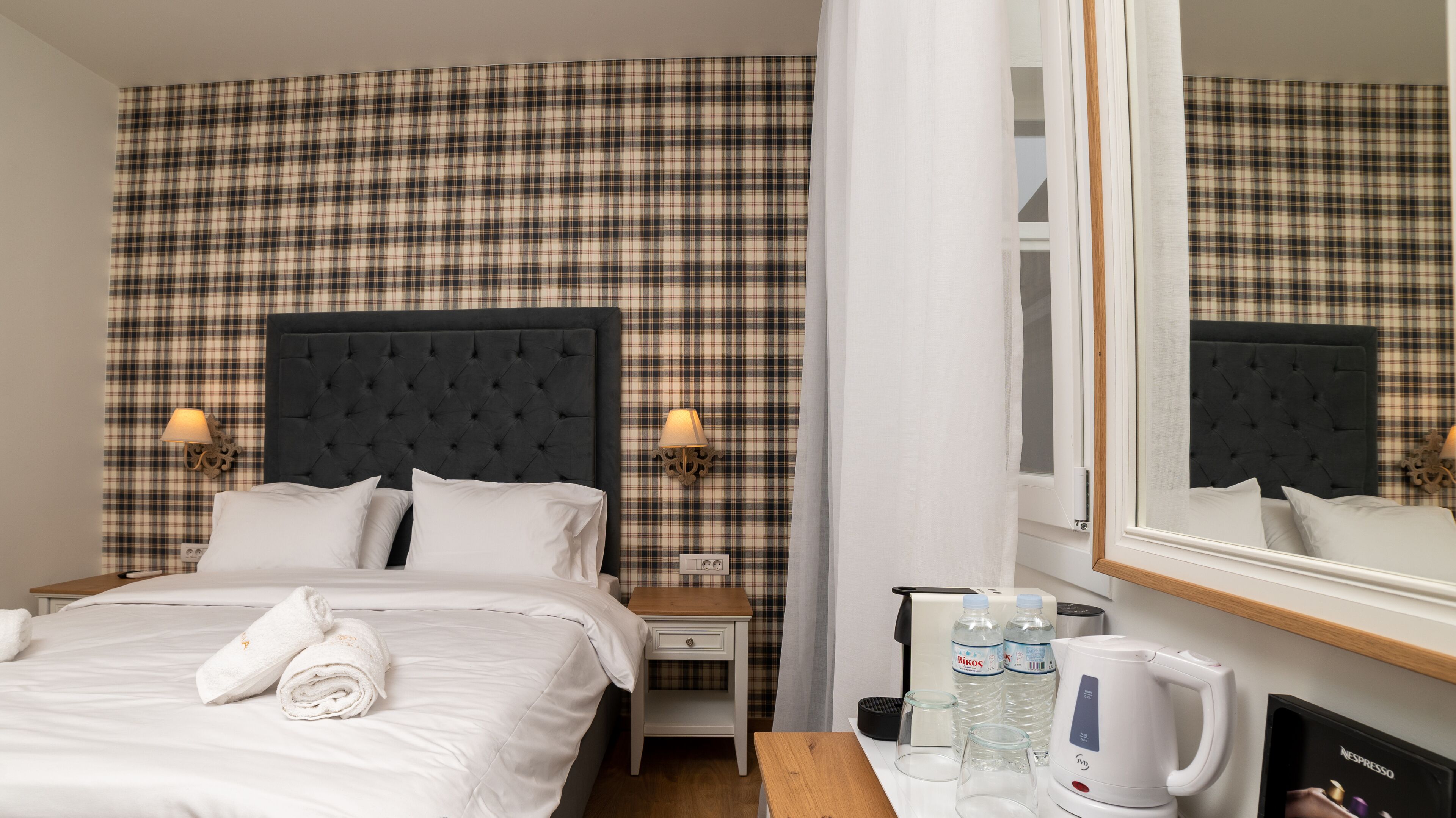 A hotel room with a plaid-patterned headboard, two bedside lamps, a bed with white linens and towels, a nightstand with a lamp, a coffee maker, and a mirror.