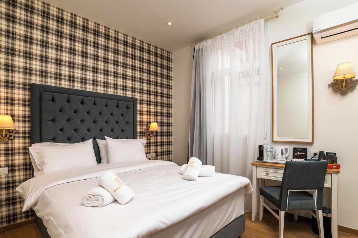 A hotel room with a plaid wallpaper, a tufted headboard, two bedside lamps, a desk with a chair, a mirror, and a bed with white linens and towels.