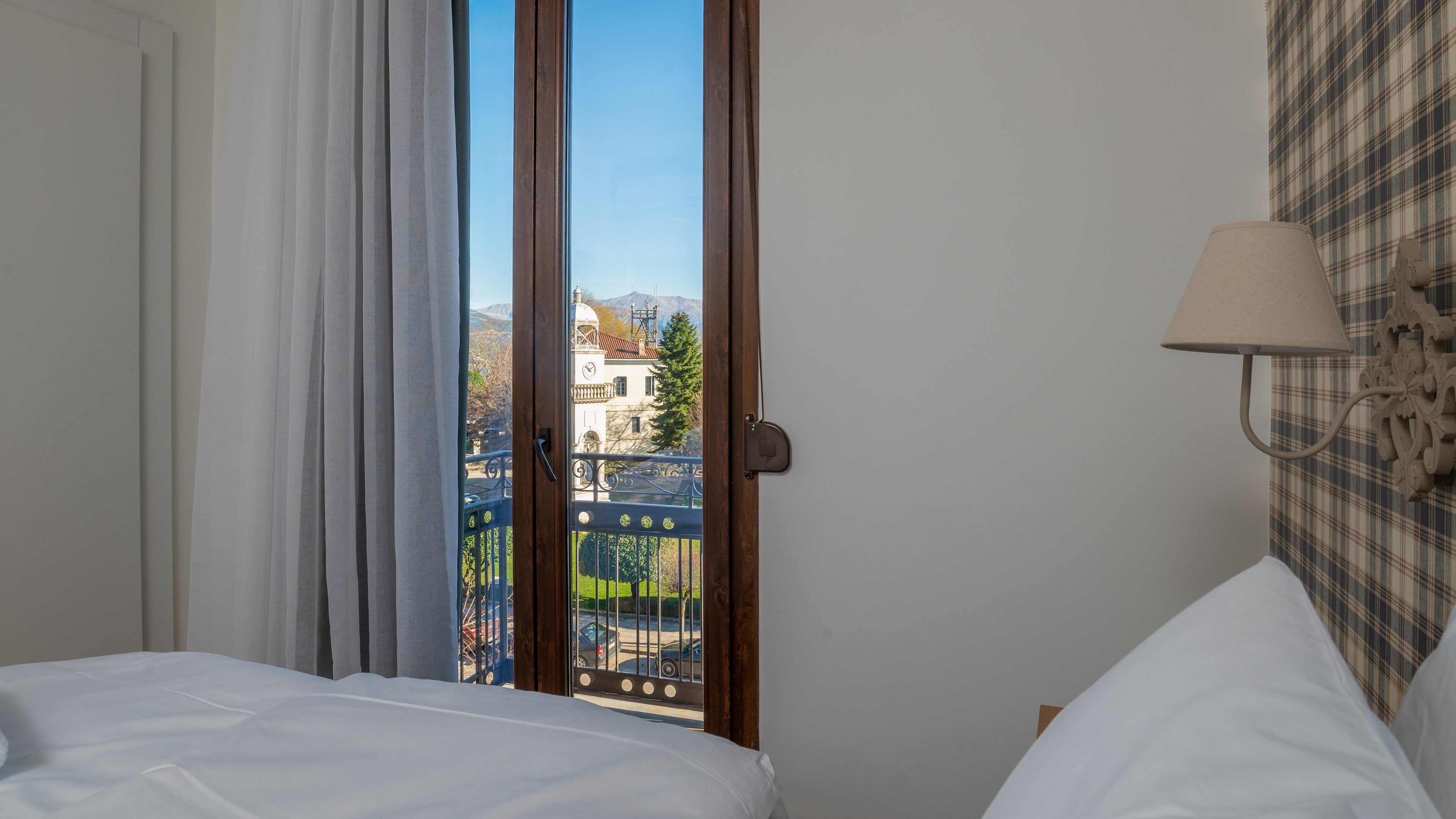 Una habitación de hotel con una cama, un ventanal con vistas a un edificio y montañas, y una lámpara fijada a la pared.