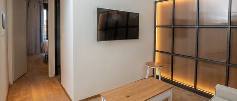 A modern hotel room with a wooden floor, a small wooden table, a white sofa, a wall-mounted TV, and a sliding door leading to another room.