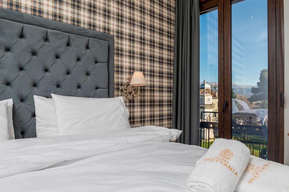A neatly made bed with white linens and a grey tufted headboard, a wall-mounted lamp, and a view of a cityscape through a large window.