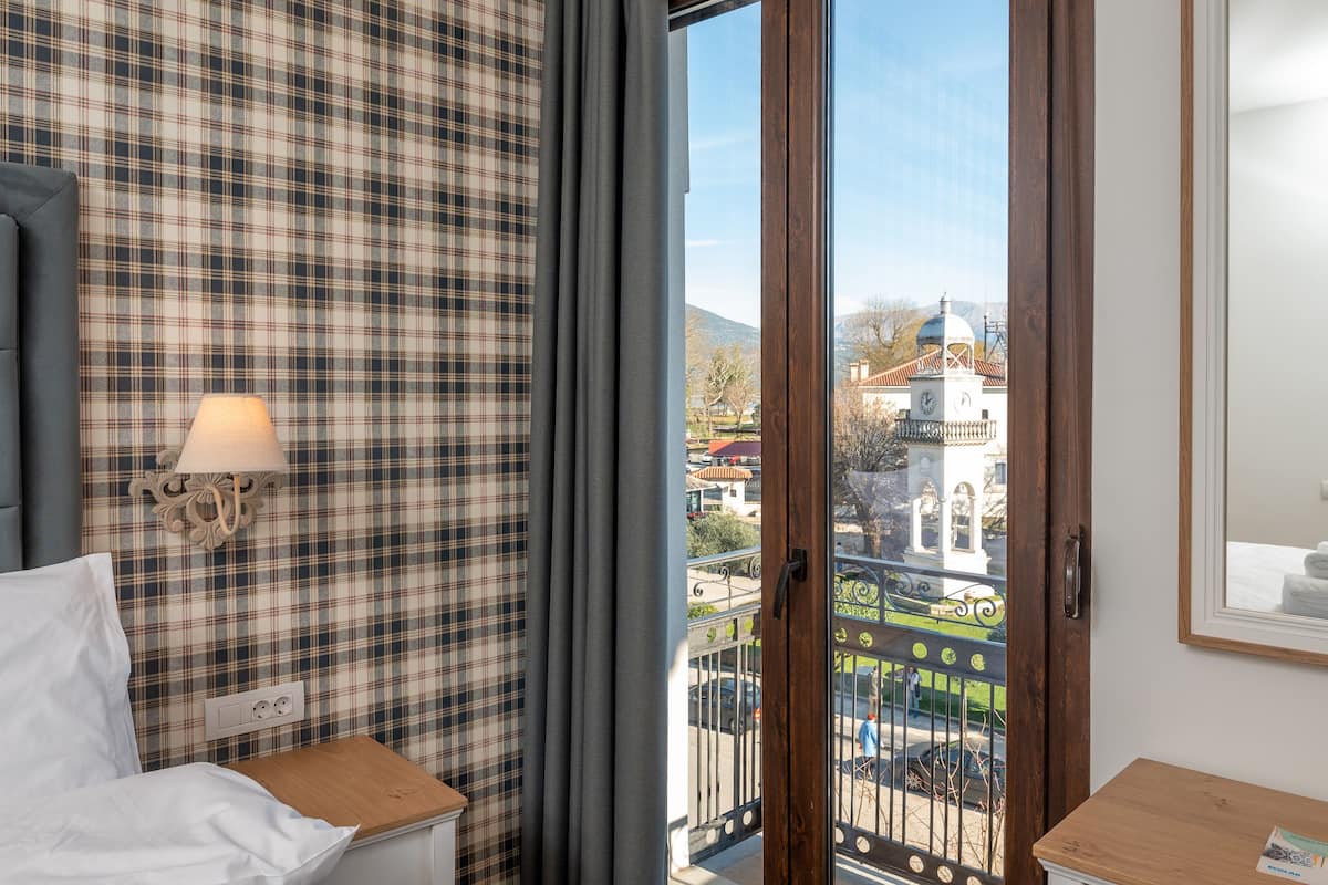 A hotel room with a plaid-patterned wall, a tufted headboard, a bedside table with a lamp, and a view of a cityscape through a glass door.