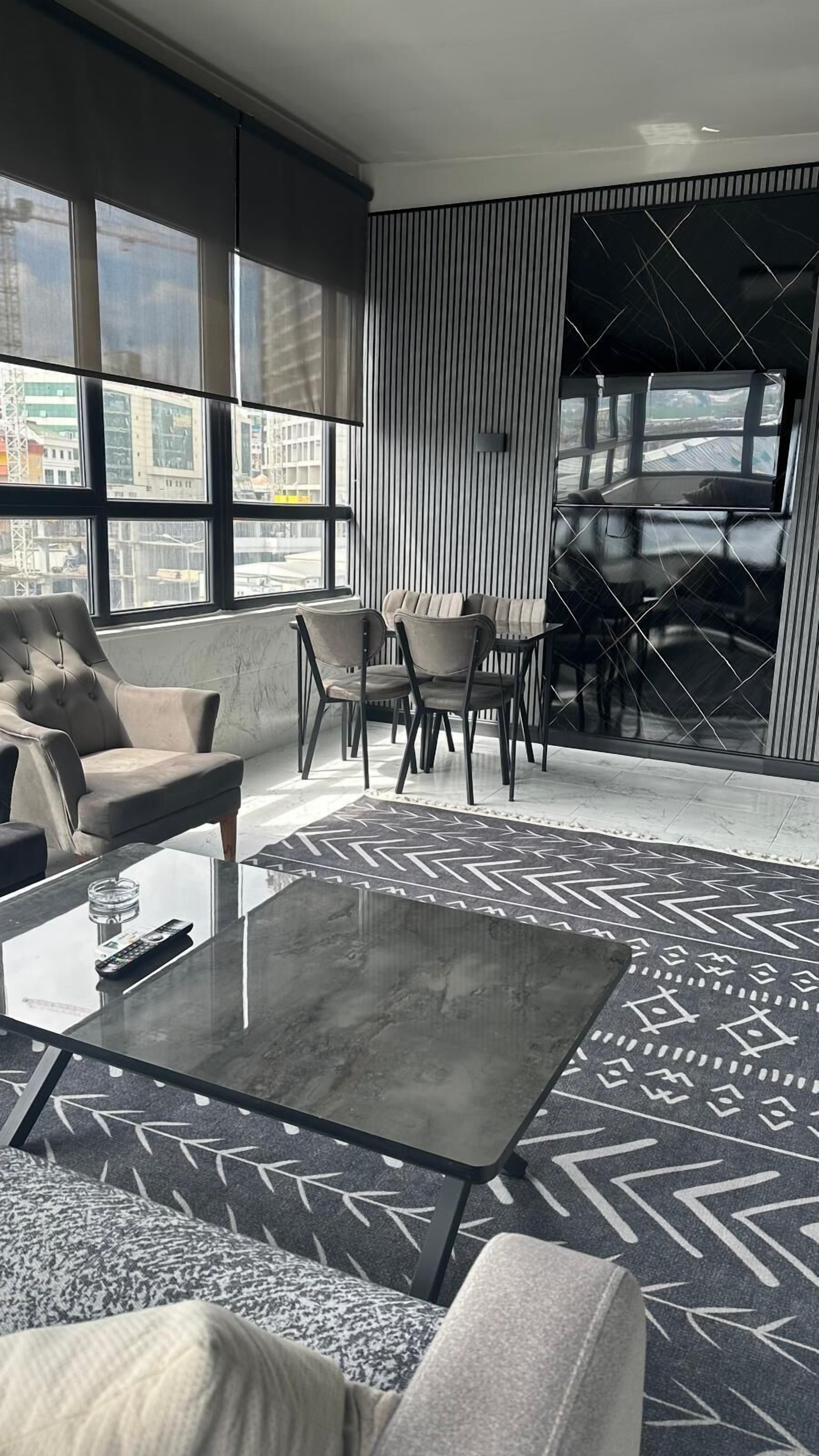 A modern hotel room with a large window, a sofa, a coffee table, and a dining area with chairs and a table.