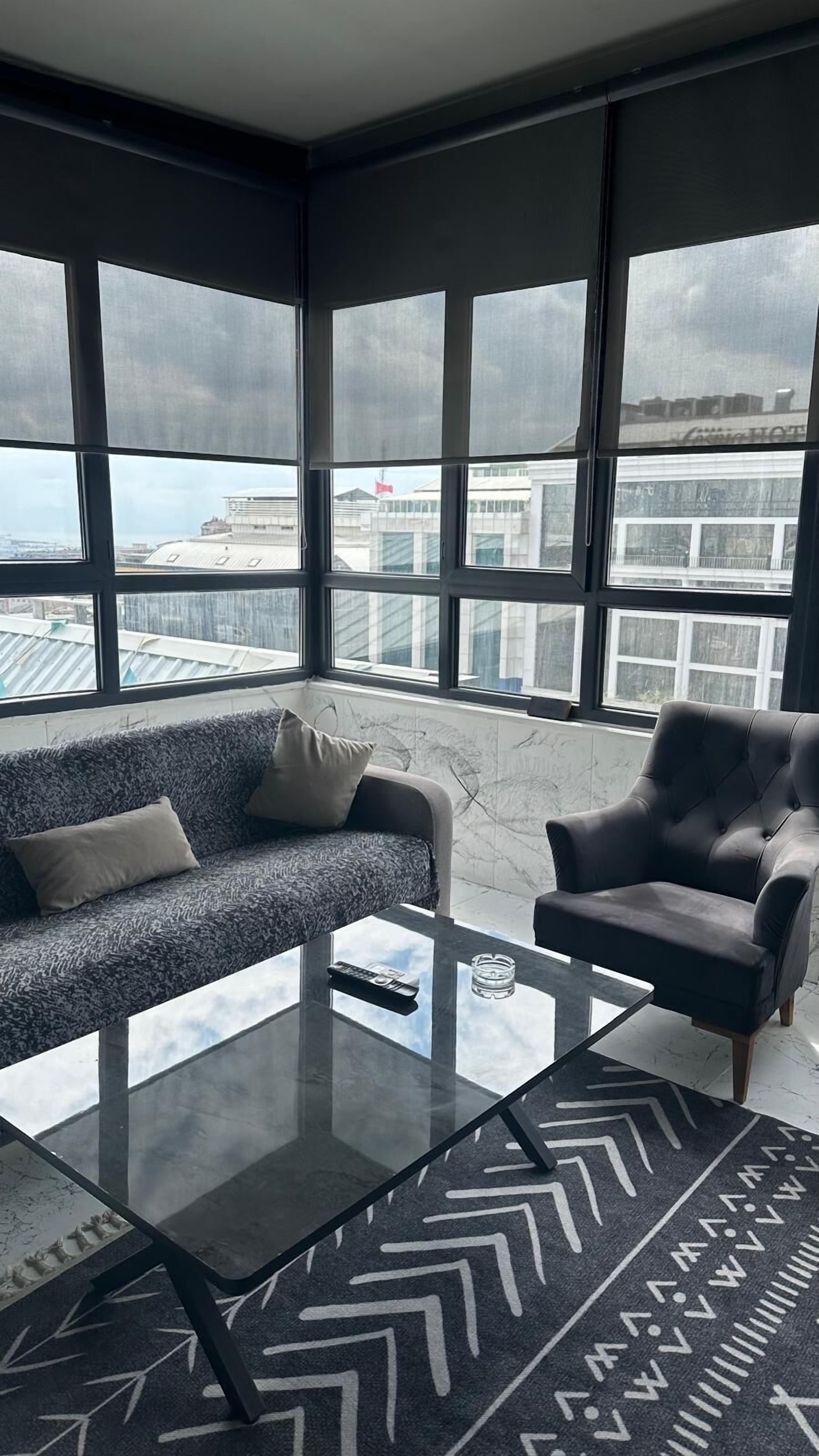 A modern living room with a sofa, armchair, and glass coffee table. Large windows offer a view of the cityscape.