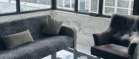 A modern living room with a sofa, armchair, and glass coffee table. Large windows offer a view of the cityscape.
