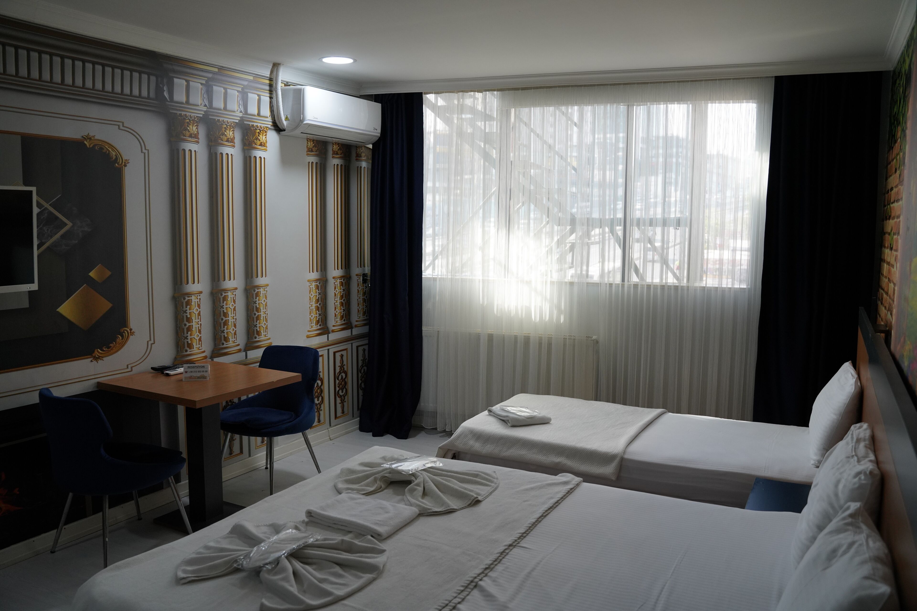 A hotel room with two beds, a desk, a chair, and a large window with sheer curtains.