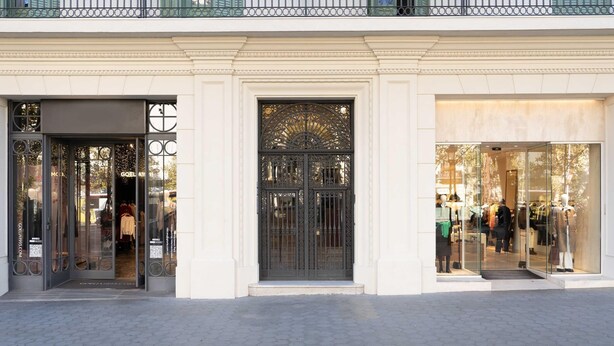 Un edificio de fachada clásica, con una entrada central adornada con ornamentación metálica y un escaparate de vidrio que muestra maniquíes.
