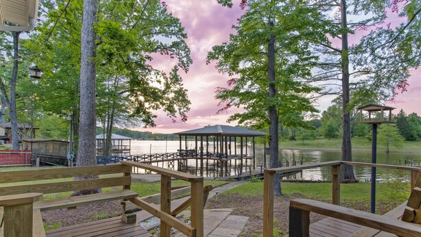 Una terraza de madera con bancos que da a un lago, un pabellón y un muelle con un bote.