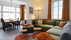 Ein modernes Wohnzimmer mit einem grünen Sofa, orangefarbenen Kissen, einem Esstisch mit Stühlen und einer Lampe.