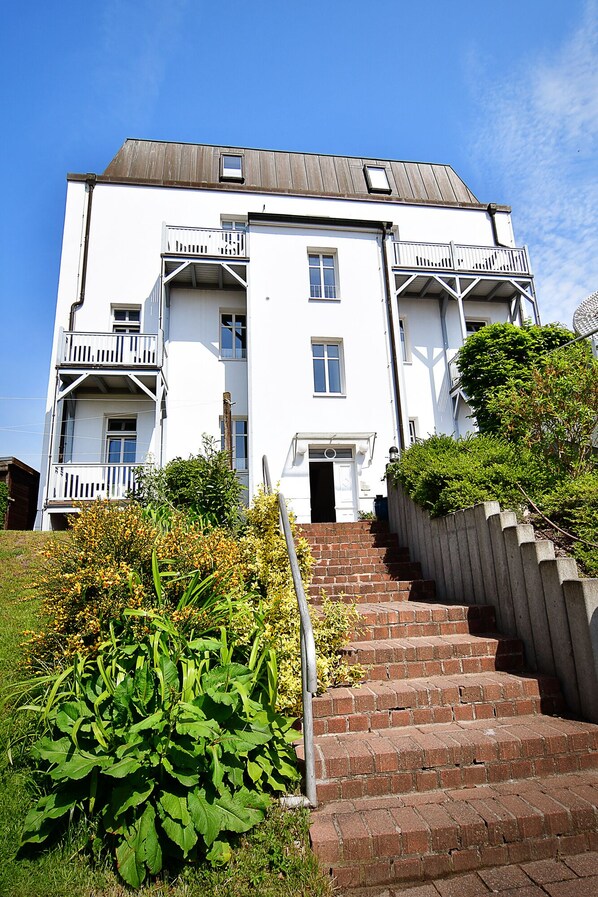 A white multi-story building with a central entrance, a small balcony on the ground floor, and a brick staircase leading up to the entrance. - Villa Hamburg Appartement 08 (Seebad Ahlbeck)