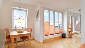 A spacious living area with a wooden floor, a dining table with chairs, large windows, and a view of the outside. - Villa Hamburg Appartement 08 (Seebad Ahlbeck)