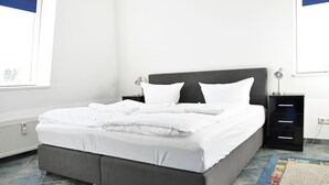 Uma cama bem arrumada com lençóis brancos, cabeceira escura e mesa de cabeceira com abajur.
