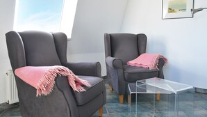 Duas poltronas cinzentas com mantas cor-de-rosa, uma mesa de centro de vidro transparente e um quadro na parede.
