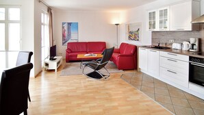 Ein gemütlicher Wohnraum mit einem roten Sofa, einem Essbereich mit Tisch und Stühlen, einer Küchenzeile inklusive Herd und Kaffeemaschine sowie Kunstwerken an den Wänden.