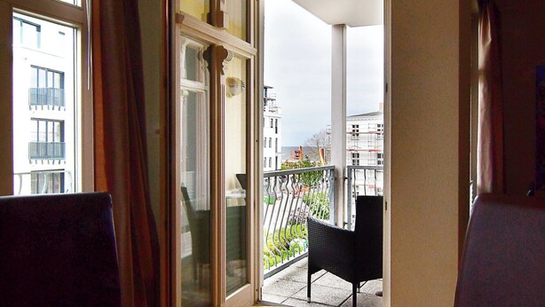 Ein Zimmer mit einem Holztisch, einem Teekessel und einer Tischdecke mit Muster. Ein Fenster mit Vorhängen, eine Tür, die nach draußen führt, und ein Blick auf Gebäude sowie ein Balkon sind zu sehen.