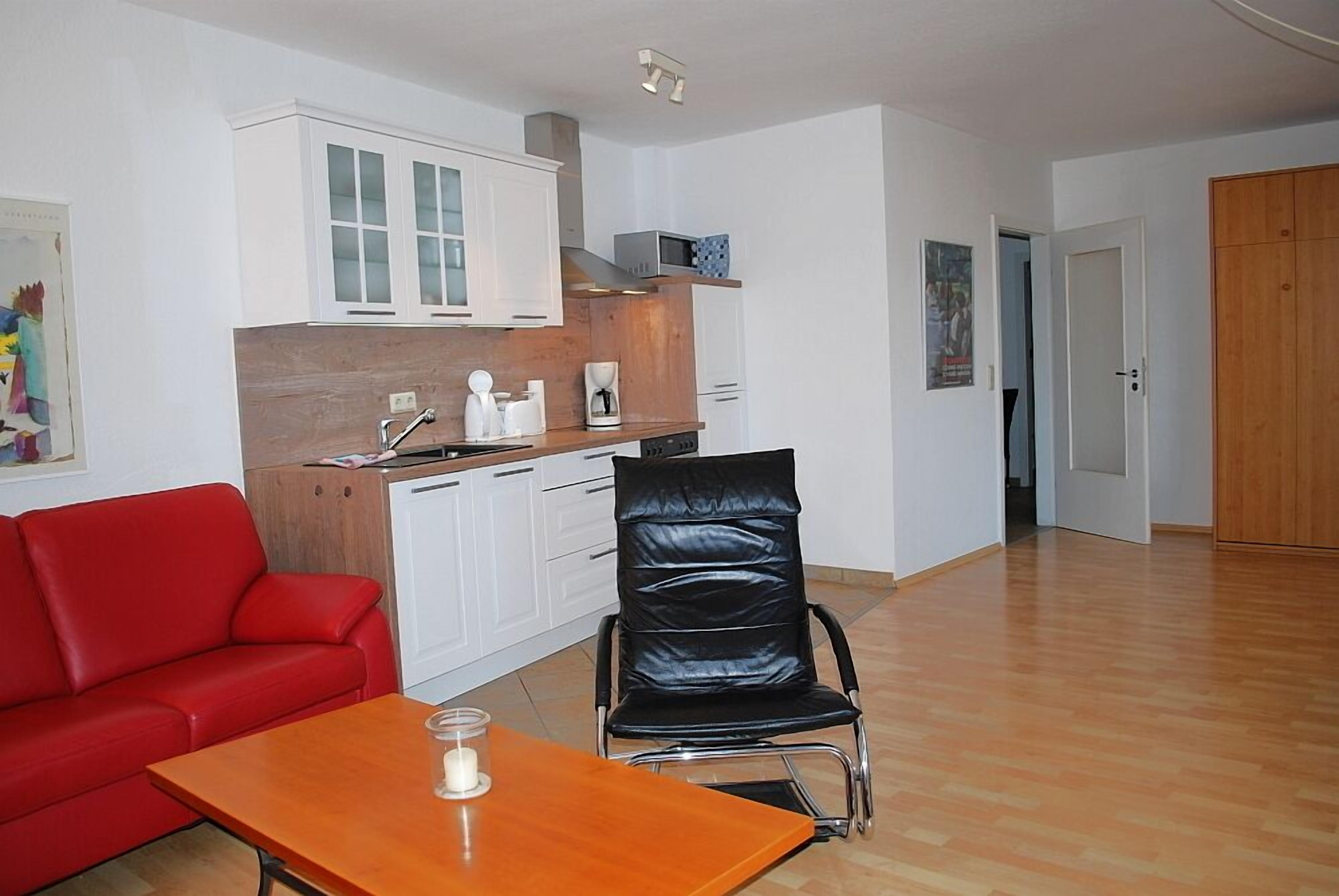 Un espace de vie compact comprenant un coin cuisine, un canapé rouge, un fauteuil en cuir noir et une table en bois.