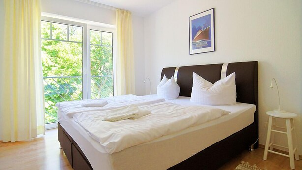 Ein Schlafzimmer mit einem großen Bett, einem kleinen Hocker und einem Fenster mit Vorhängen.