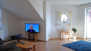 Ein Wohnzimmer mit Fernseher, einem kleinen Tisch mit Flasche und Blumen, einem blauen Teppich, einem Holzstuhl und einer Topfpflanze.