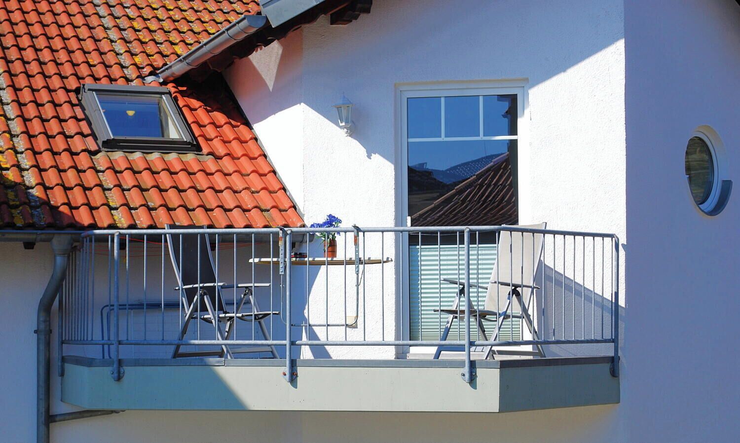 Un bâtiment à deux étages, doté d’un toit recouvert de tuiles rouges, d’un balcon avec des balustrades en métal et d’une petite fenêtre.