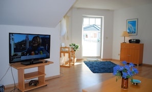 Sala de estar com televisão, móveis de madeira, tapete azul e um vaso com flores.