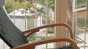 En træstol med sort vævet sæde og ryglæn, placeret foran et stort vindue med udsigt til hav og grønne omgivelser.