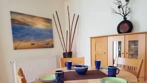 Et spiseområde med et rundt træbord, blå og grønne tallerkener samt matchende blå krus. På væggen hænger et maleri, og der er en radiator samt et skab med glasdøre.