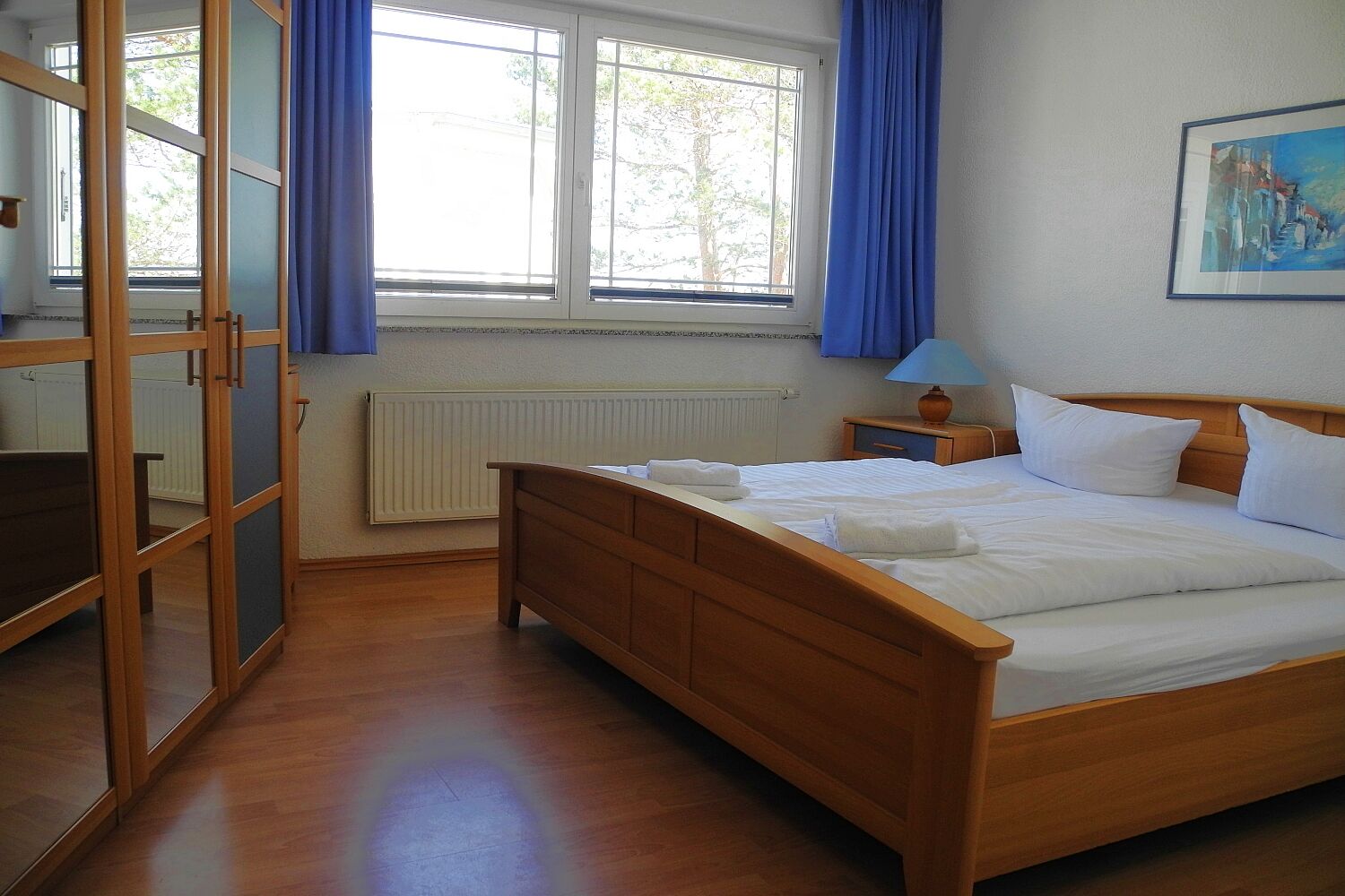 A bedroom with a wooden bed, a nightstand with a lamp, a radiator, and a window with blue curtains.