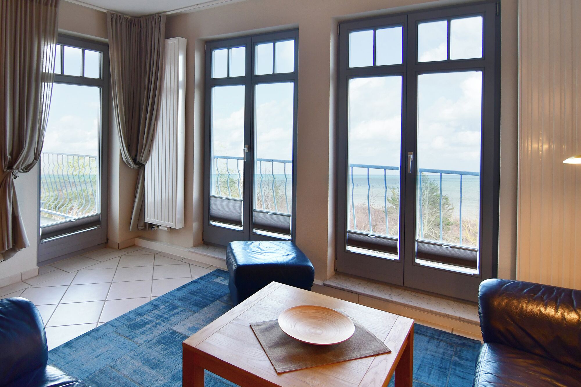 Ein Zimmer mit großem Fenster, einem Holztisch mit Schüssel, einem blauen Pouf und einem schwarzen Ledersofa.