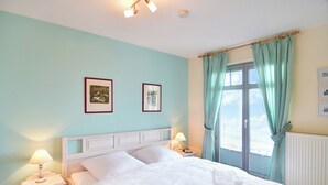 Un dormitorio con una cama, dos mesitas de noche con lámparas, un ventanal con cortinas y dos cuadros enmarcados en la pared.