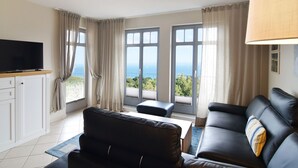 Una sala de estar moderna con televisor, dos sofás, una mesa de centro y ventanas amplias con vista al mar.