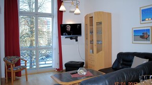A living room with a TV, a wooden cabinet, a sofa, a chair, and a window with red curtains. - Seeschloss Appartement 05 (Seebad Heringsdorf)