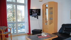 A living room with a black leather sofa, a wooden cabinet with glass doors, a television mounted on the wall, and a window with red curtains. - Seeschloss Appartement 05 (Seebad Heringsdorf)